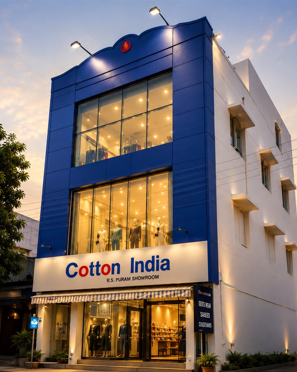The Cotton India showroom in R.S. Puram at dusk — three lit floors behind a blue glass facade, mannequins visible in every window